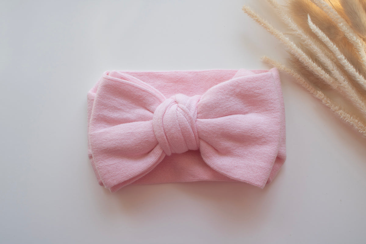 A light pink fabric headband with a large bow, laid flat on a white surface with dried grass stems in the top right corner.