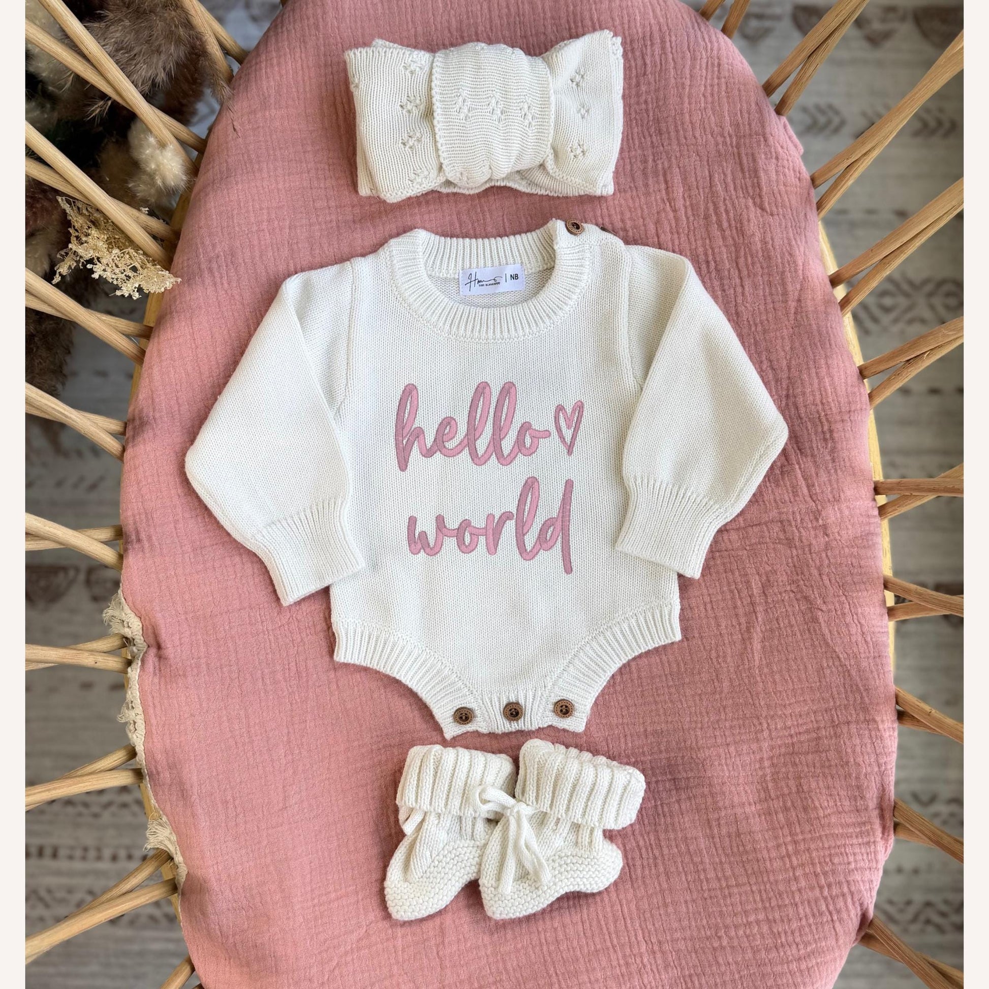 Baby outfit set with 'hello world' romper, headband, and booties on a pink surface.
