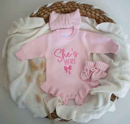 Pink baby outfit with 'She's Here' text and bow, including hat, and socks.