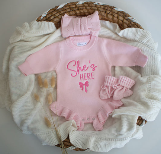 Pink baby outfit with 'She's Here' text and bow, including hat, and socks.